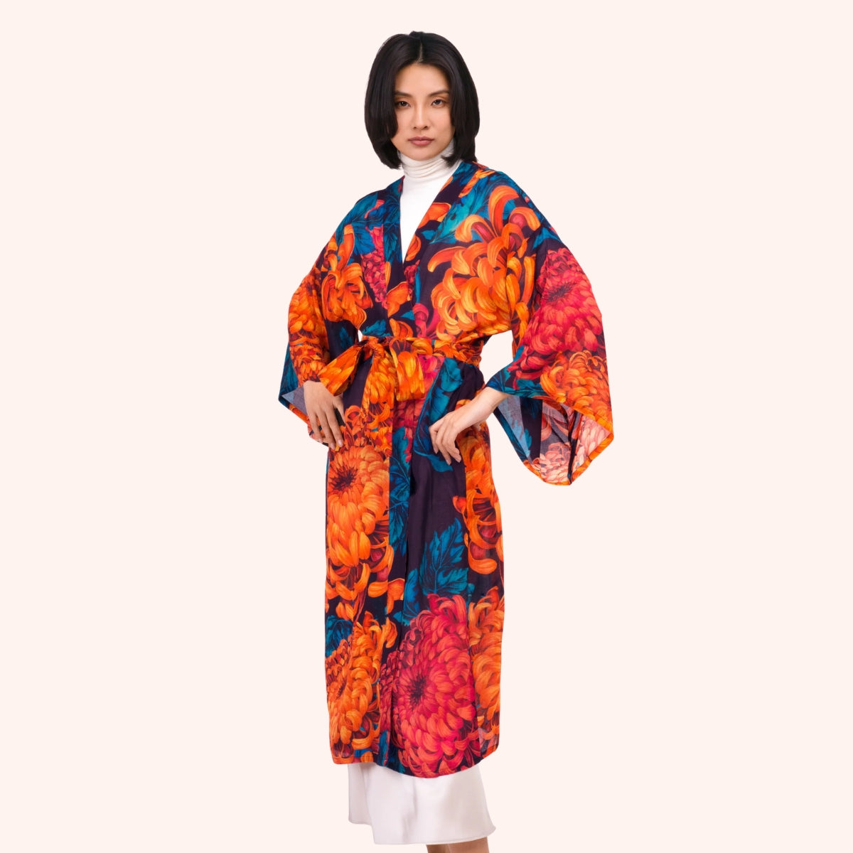 Model wearing a floor length orange and teal chrysanthemum print kimono gown with waist tie, over a white turtleneck
