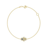 A delicate gold chain bracelet with a Hamsa hand charm paved with small clear stones and featuring a central blue gemstone, shown on a plain white background.