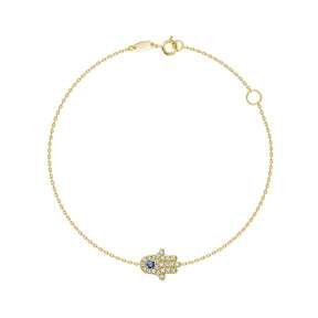 A delicate gold chain bracelet with a Hamsa hand charm paved with small clear stones and featuring a central blue gemstone, shown on a plain white background.