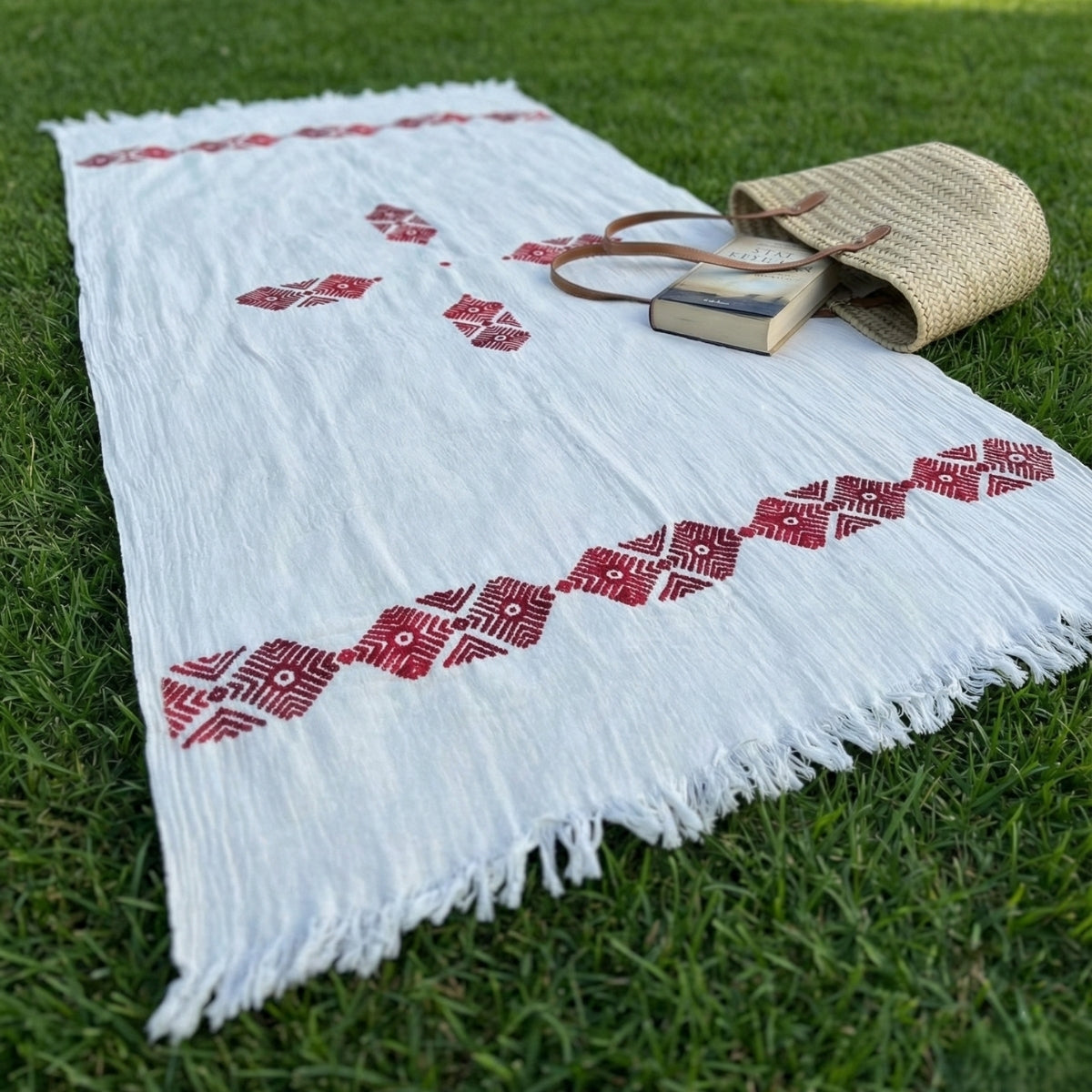 White shawl with red patterns on grass, accompanied by a straw bag and book.