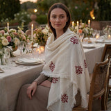 Woman sitting at a decorated outdoor dinner table with floral arrangements and candles wearing a white shawl with intricated printed patterns.