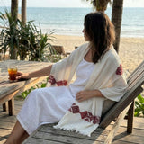Woman sitting on a wooden deck by the beach, holding a drink, with a white dress and patterned shawl.