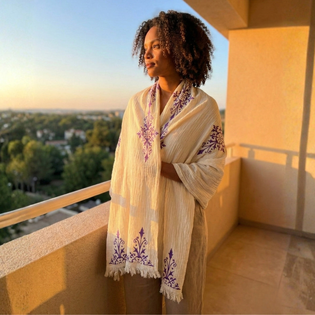 Woman standing on a balcony wearing a beige embroidered scarf with a scenic view in the background.