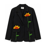 Flat front view of the Boutique Flo Sunflower Bloom Embroidered Black Jacket, showing hand-embroidered sunflowers with green stems across the black denim, notched lapel collar, and flap pockets