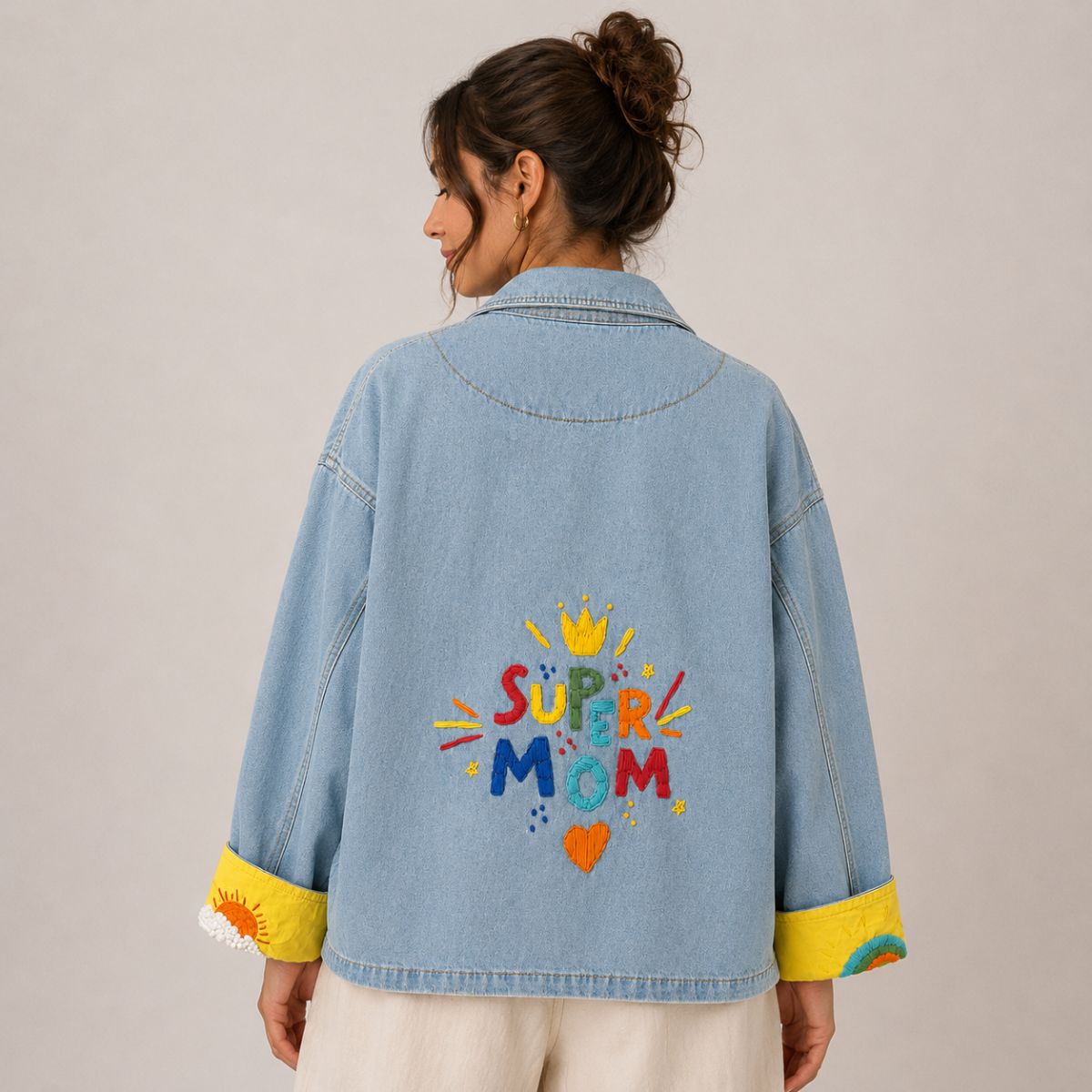 Back view of the Boutique Flo Super Mom Embroidered Light Denim Jacket worn by a model, showing the multicolor hand-embroidered "Super Mom" lettering with yellow crown and orange heart on light-wash denim