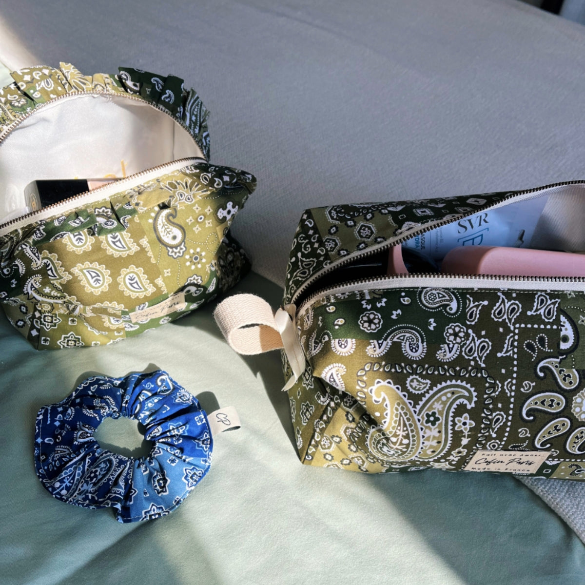 Lifestyle shot featuring green bandana print toiletry bags and a matching blue bandana hair scrunchie on a bed.