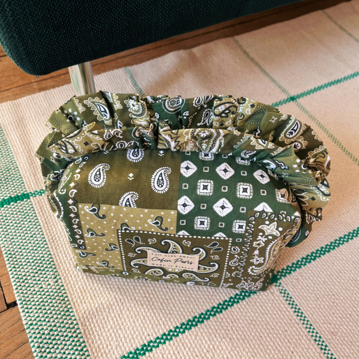 Close-up lifestyle shot of the Cofin Paris Medium Louise Green Bandana Ruffled Toiletry Bag resting on a woven rug.
