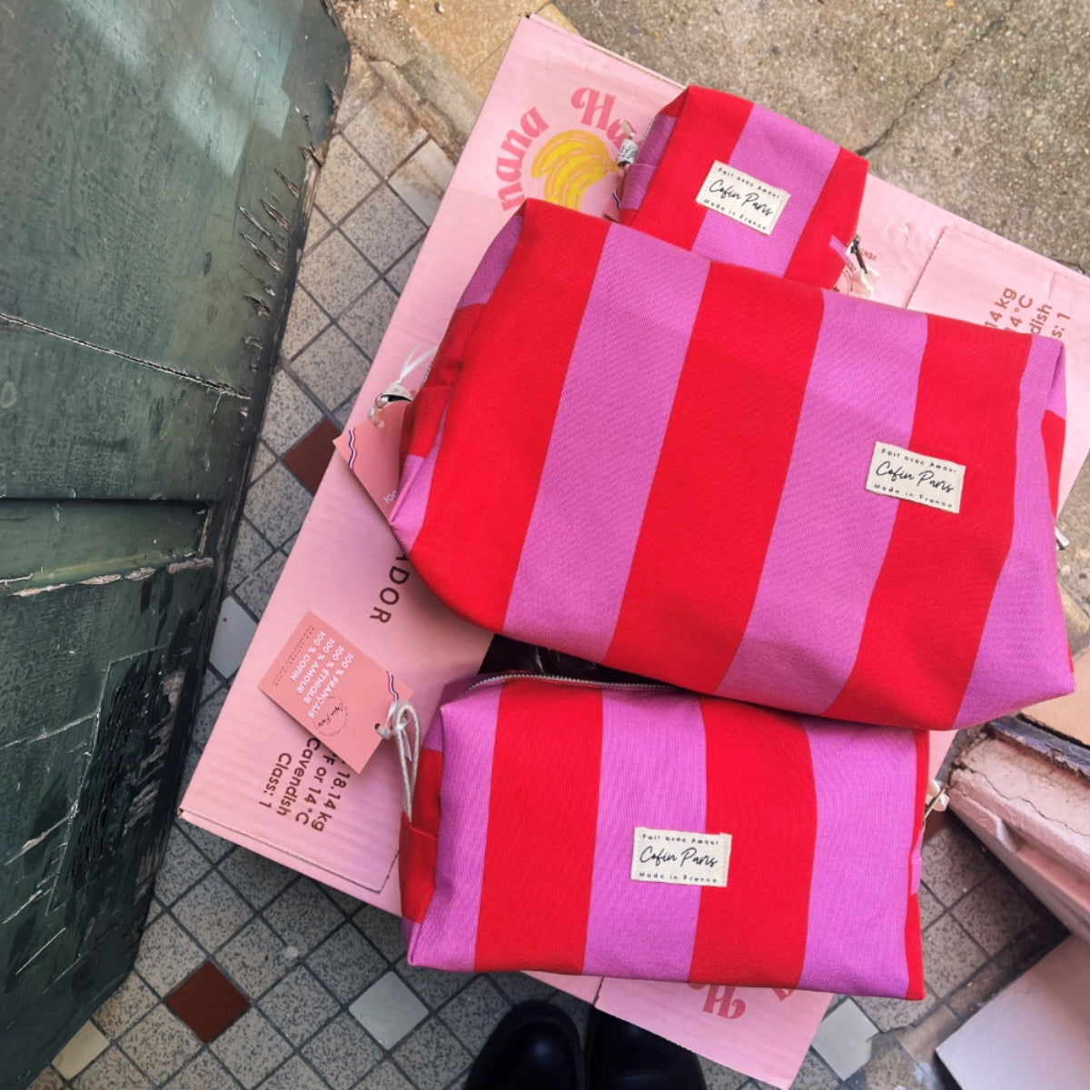 Top-down view of three Cofin Paris red and pink striped toiletry pouches arranged on pink boxes, featuring the XS Paola Striped pouch at the top.