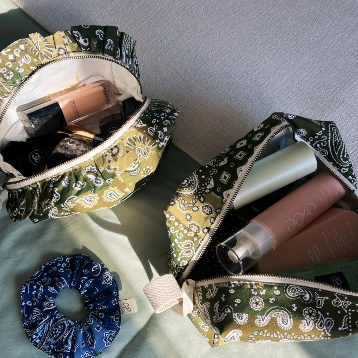 Overhead view of two open Cofin Paris green bandana toiletry bags filled with beauty products and cosmetics next to a blue bandana scrunchie.