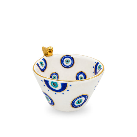 Front view of a white ceramic bowl decorated with hand-painted blue evil eye patterns, a metallic gold rim, and a small gold bird perched on the edge.