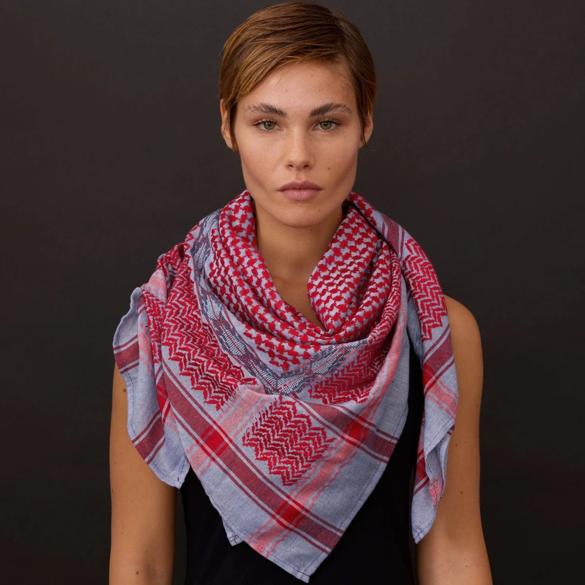 Front view portrait of short-haired model wearing SEP blue and red keffiyeh scarf layered around her neck against a dark background