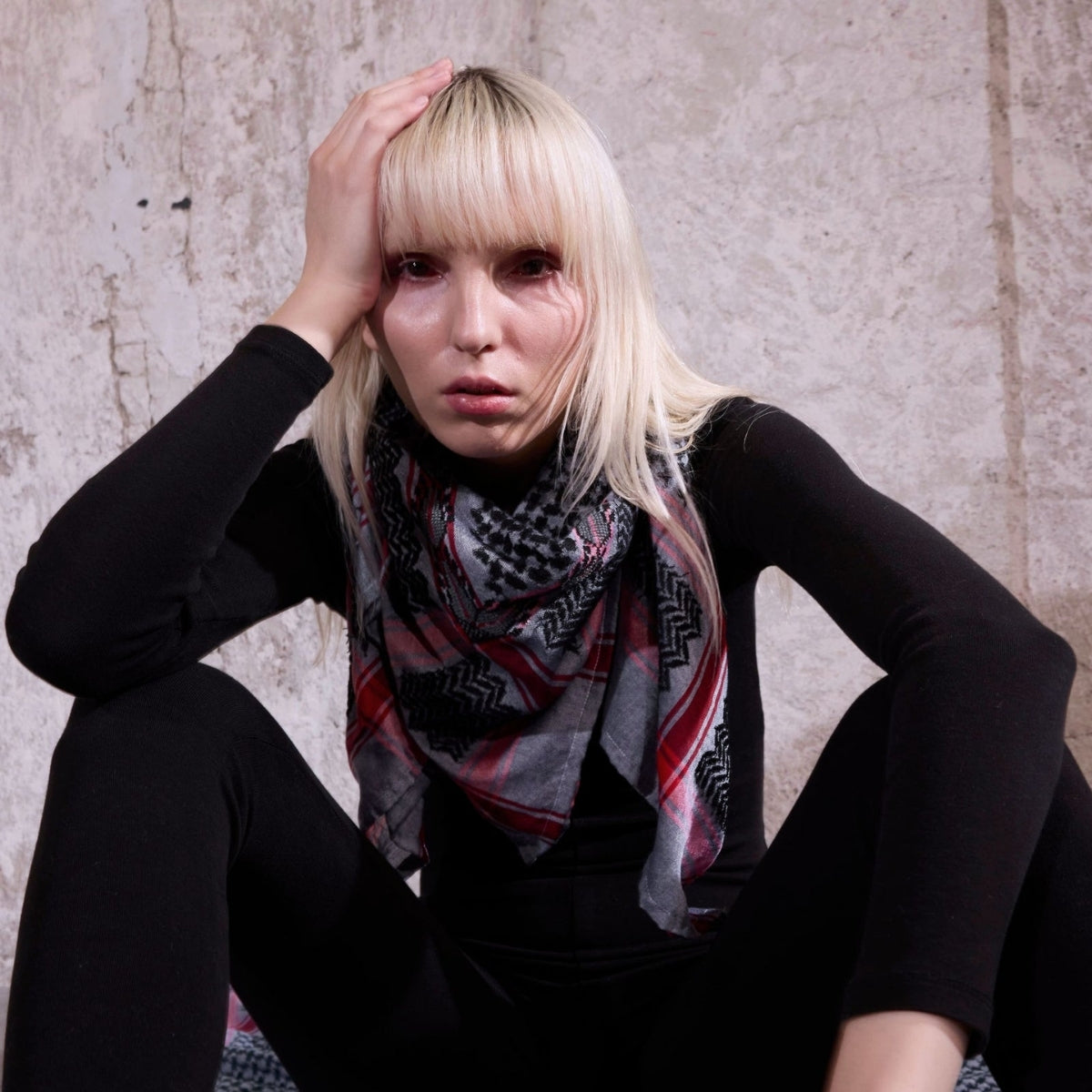 Model wearing SEP grey and black keffiyeh scarf layered around her neck sitting against a textured wall