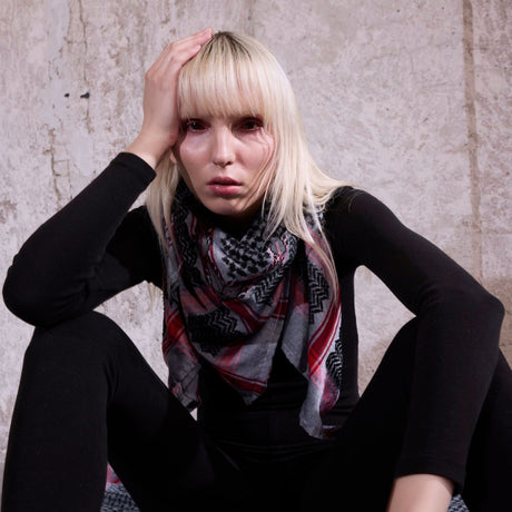 Model wearing SEP grey and black keffiyeh scarf layered around her neck sitting against a textured wall