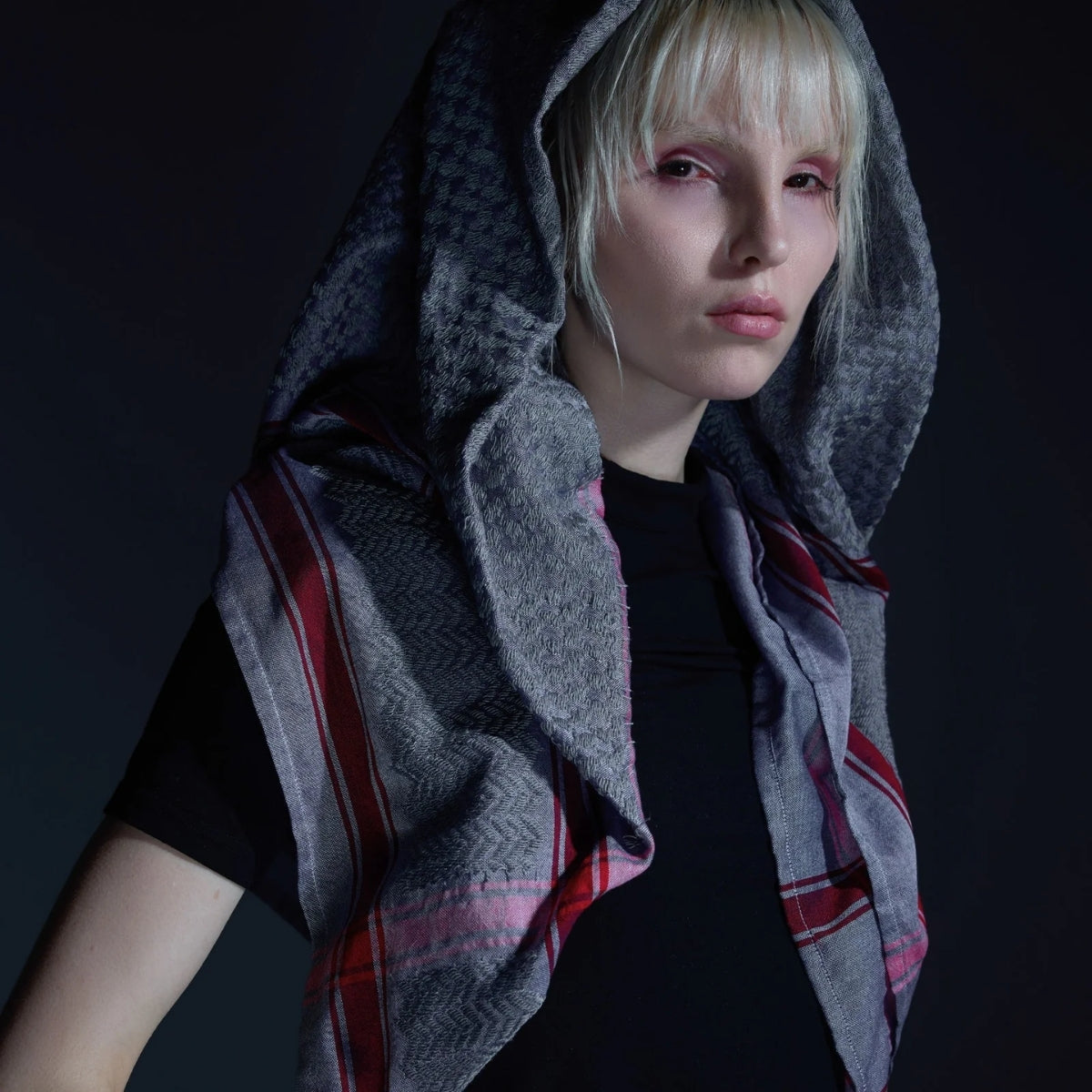Dramatic studio portrait of model wearing SEP grey thalia keffiyeh scarf styled over her head like a hood against a dark background