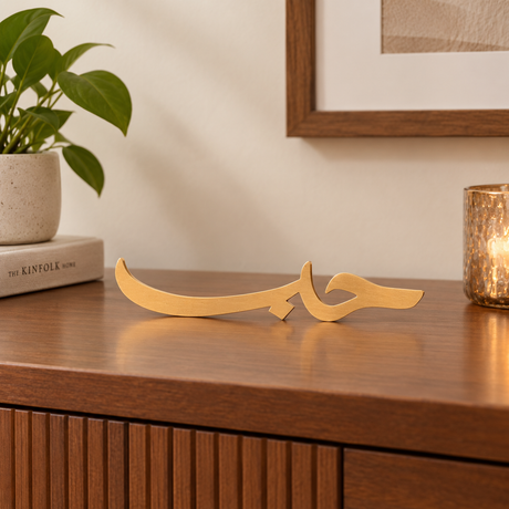Tabletop view of the You & Eye Arabic Calligraphy Love copper metal decor displayed on a wooden credenza next to a potted plant and candle.